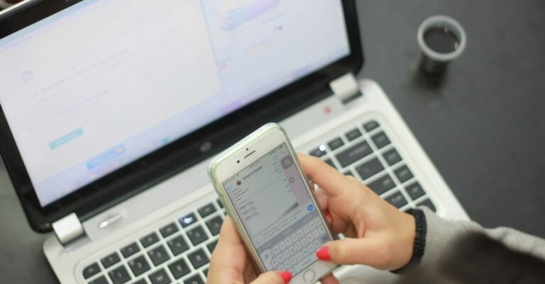 Close-up of woman texting on a smartphone while a laptop is open, indicating multitasking.