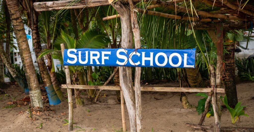 A rustic surf school sign among palm trees on a sandy tropical beach.